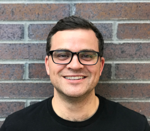 Picture of Jacob Fowler from the shoulders up facing the camera smiling wearing a black tee shirt and black glasses